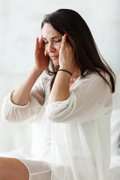 中年妇女坐在家中卧室的一张白色床上，双手触摸着头，同时感到头痛、疼痛和不适。因失眠或偏头痛而痛苦 — 图库照片