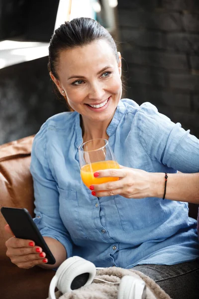 Leisure time concept. Happy beautiful woman talks on a phone and drinks orange juice from glass sitting on a couch indoors. Female spending her free day and relaxing at home alone