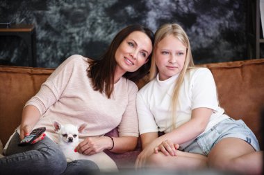Anne-kız ilişkisi kavramı. Bir kadın ve genç bir kız kanepede TV karşısında oturmuş eğleniyorlar. Aile evde zaman geçirir. Küçük köpek kucağında yatıyor.
