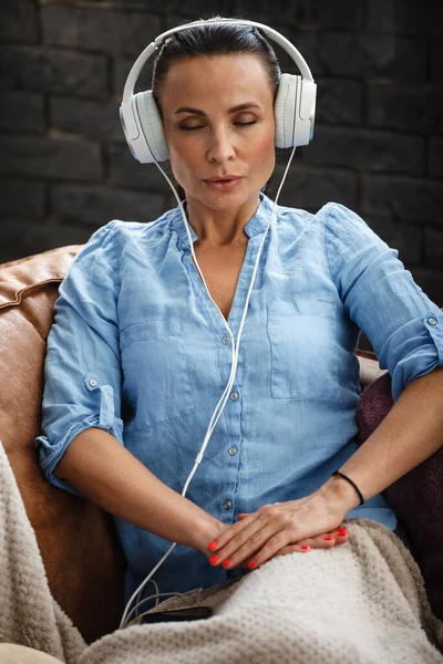 Vrije tijd concept. Gelukkig mooie vrouw luisteren naar de muziek met behulp van koptelefoon zitten op een bank binnen. Vrouw brengt haar vrije dag door en ontspant alleen thuis. — Stockfoto