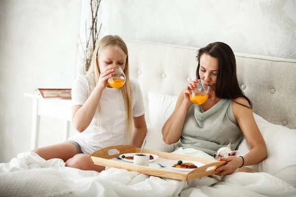 ベッドのコンセプトで家族の朝食を持っています 若い幸せな母親はパンケーキを食べ 10代の娘とオレンジジュースを飲む 女性と女の子は朝寝室に座って — ストック写真