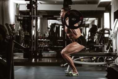 Spor, spor ve sağlıklı yaşam tarzı konsepti. Çıplak gövdeli yakışıklı atletik adam egzersiz yapıyor. Vücut geliştiren erkek model kaslar halterle çömelme hareketleri yapıyor. Yan görünüm