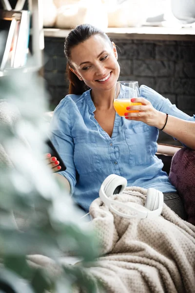 Έννοια ελεύθερου χρόνου. Ευτυχισμένη όμορφη γυναίκα μιλάει σε ένα τηλέφωνο και πίνει χυμό πορτοκάλι από γυαλί που κάθεται σε έναν καναπέ σε εσωτερικούς χώρους. Γυναίκα ξοδεύει την ελεύθερη ημέρα της και χαλαρώνει στο σπίτι μόνη της. — Φωτογραφία Αρχείου