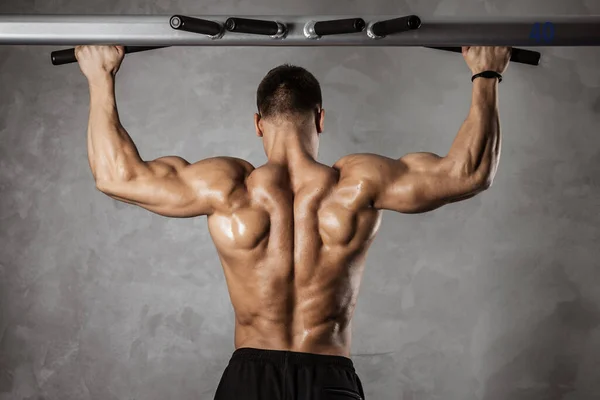 Fitness en gimnasio, deporte y estilo de vida saludable. Hombre atlético guapo con torso desnudo haciendo ejercicios. Culturista modelo masculino bombeo de los músculos haciendo tira hacia arriba en el travesaño. Vista trasera — Foto de Stock