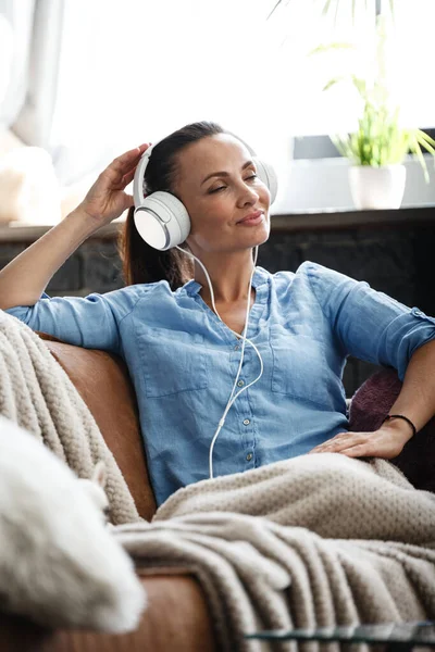 Boş zaman konsepti. Kapalı bir koltukta oturmuş kulaklık takarak müzik dinleyen mutlu güzel bir kadın. Kadın boş gününü evde yalnız geçirir.. — Stok fotoğraf