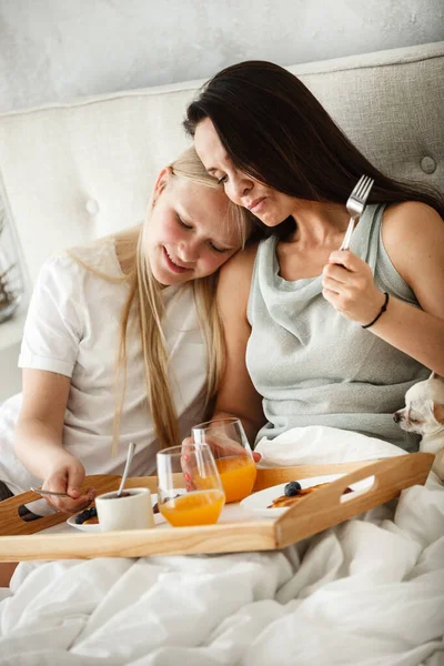 침대에서 아침 식사를 하는 거죠. 행복 한 어머니는 팬케이크를 먹고 십 대 딸과 함께 오렌지 주스를 마십니다. 아침에 침실에 앉아 있는 여자와 여자. — 스톡 사진