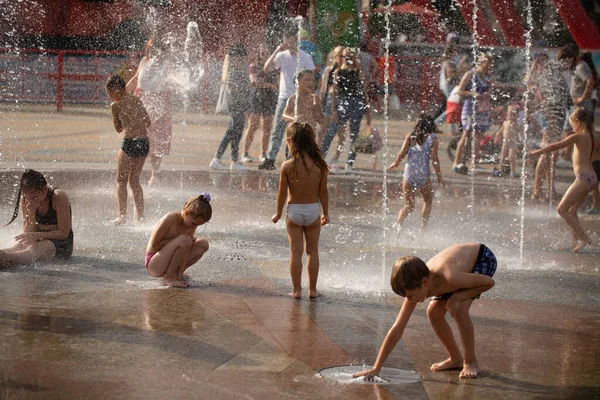 Yaz eğlencesi. Mutlu çocuklar koşar, jetlerin arasında oynar ve fıskiyeye su sıçratır. Şehir parkında yaz günü. Kharkiv, Ukrayna. 08.08.2021