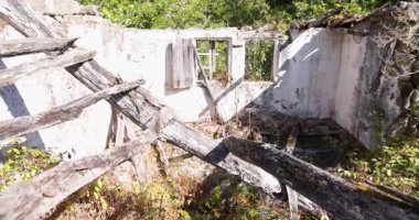 A destroyed house with a collapsed roof and broken windows. The building is dilapidated with wooden beams fallen inside the building. Tilt down shot.