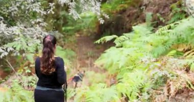 Kadının ve iki köpeğinin eğrelti otlarıyla yemyeşil bir ormanda yürürken çekilmiş bir fotoğrafı. Kadın siyah gömlek ve pantolon giyiyor..