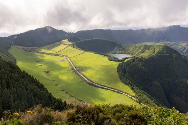 Sete Cidades, Azores 'de içinden geçen bir dağ sırası. Yol yemyeşil çimenler ve ağaçlarla çevrili. Gökyüzü bulutlu, ortama huzurlu ve huzurlu bir atmosfer veriyor.