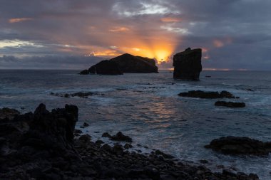 Okyanus sakin ve gökyüzü turuncu. Güneş kayaların arkasında batıyor. Su mavi ve kayalar gri.