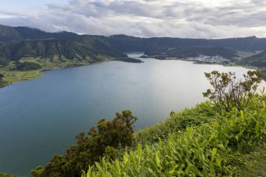 Sete Cidades, Azores 'in arka planında yeşil bir yamaç olan güzel bir göl. Su sakin ve berrak. Gökyüzü bulutlu, ama güneş bulutların arasından parlıyor.