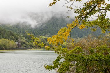 Arka planda bulutlu bir gökyüzü olan bir göl ve kilise. Ağaçlar yeşil ve yapraklar sarı Nossa Senhora das Vitorias, Furnas