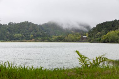 Arka planda kilise olan bir göl. Gökyüzü bulutlu ve su sakin. Nossa Senhora das Vitorias, Furnas 'ta manzara huzurlu ve huzurlu.
