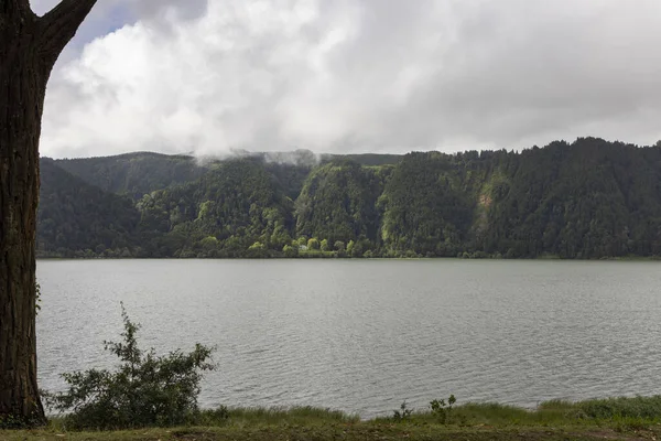 Önünde ağaç olan büyük bir su kütlesi. Gökyüzü bulutlu ve Nossa 'da su sakin Senhora das Vitorias, Furnas