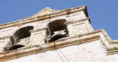 The Romanesque church bell tower is old and has a lot of moss growing on it. The sky is clear and blue