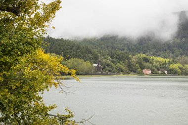 Arkasında bulutlu bir gökyüzü olan bir göl. Ağaçlar yeşil ve sarı. Nossa Senhora das Vitorias, Furnas 'ta su sakin ve berrak.