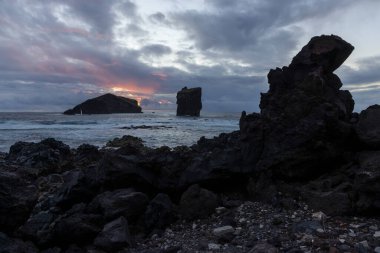 Önünde büyük bir kaya olan kayalık bir kıyı şeridi. Gökyüzü bulutlu ve güneş batıyor.