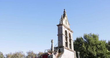 Çan kulesi olan uzun, eski bir kilise kulesi. Gökyüzü açık ve mavi. Kule ağaçlarla çevrili.