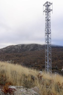 Bir tarlanın ortasında uzun metal bir kule duruyor. Gökyüzü bulutlu ve çimenler kuru. Kule ağaçlarla çevrili ve manzara huzur dolu.