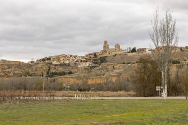 Önünde bir kilise ve ağaç olan küçük bir kasaba. Gökyüzü bulutlu ve şehir tepelerle çevrili.