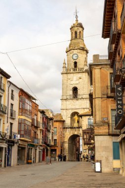 Toro, İspanya, 10 Aralık 2025, ortasında saat kulesi olan bir şehir sokağı. Saat kulesi uzun ve üzerinde bir saat var. Bölgede dolaşan bir sürü insan var..