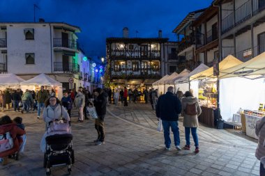Candeleda, Avila, İspanya, 10 Aralık 2025: Kalabalık bir cadde, insanların yürüdüğü ve bebek arabasında bir bebek var. Sokak ışıklarla aydınlatılmış ve satılık çadırlar var..