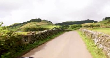 Yan tarafında taş duvar, arka tarafında da Sao Miguel Azores tepesi olan bir yol. Yol boş ve gökyüzü bulutlu. İzleme görüntüsü.
