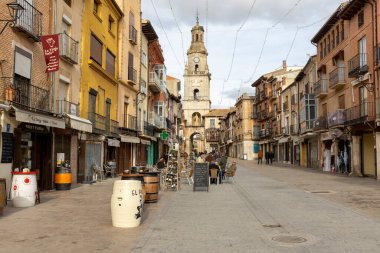 Toro, İspanya, 10 Aralık 2025: Arkasında saat kulesi olan işlek bir şehir caddesi. Caddede birkaç kafe ve restoran var ve insanlar etrafta dolanıyor.