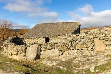 Sazdan çatılı küçük taştan bir ev bir tarlada oturur. Ev kayalarla çevrili ve önünde büyük bir kaya var. Gökyüzü açık ve güneş parlıyor.