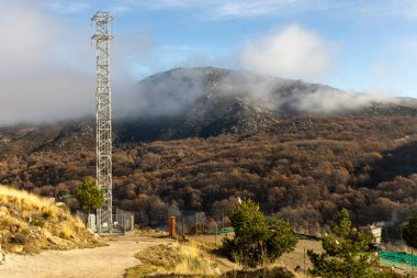 Uzun bir kule bir ormanın ortasındadır. Gökyüzü bulutlu ve ağaçlar kahverengi.