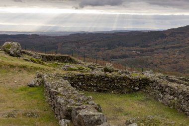 Taş bir duvarı ve çimenli bir alanı olan kayalık bir yamaç. Güneş bulutların arasından parlıyor, sıcak ve huzurlu bir atmosfer yaratıyor.