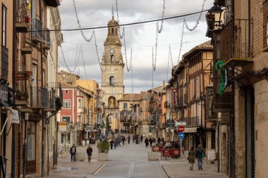 Toro, İspanya, 10 Aralık 2025: Arkasında saat kulesi olan işlek bir şehir caddesi. İnsanlar etrafta dolaşıyor ve bir sürü dükkan var.