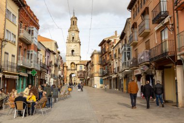 Toro, İspanya, 10 Aralık 2025: İnsanların yürüdüğü ve masalarda oturduğu yoğun bir cadde. Cadde binalarla ve saat kulesiyle dolu.