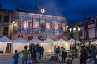 Candeleda, Avila, İspanya, 10 Aralık 2025: Yanı başında büyük bir saatle bir binanın önünde bir grup insan toplandı. Sahne ışıklarla aydınlanıyor ve insanlar ateşin etrafında dikiliyorlar.