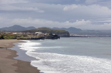 Kayalık bir kıyı şeridi ve bulutlu bir gökyüzü olan bir sahil. Kumsal boş ve dalgalar küçük.