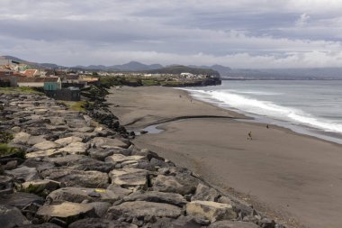 Azores, Portekiz, 8 Ekim 2025: Kayalık bir kıyı şeridi ve sahilde yürüyen birkaç kişi. Sahil kalabalık değil ve su sakin.