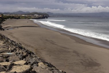 Azores, Portekiz, 8 Ekim 2025: Kayalık bir sahil ve arka planda küçük bir kasaba. Kumsal boş ve su sakin.