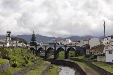 Ribeira Grande, Azores, Portekiz, 8 Ekim 2025: Tepesinde bulutlu bir gökyüzü olan bir kasabada bir nehir boyunca uzanır. Köprü binalar ve yeşil bir tepe ile çevrilidir.