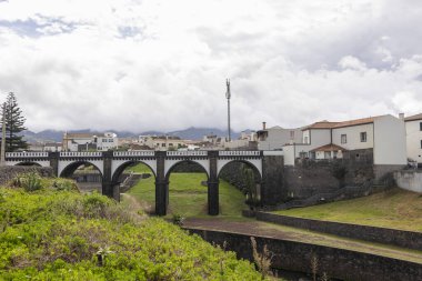 Ribeira Grande, Azores, Portekiz, 8 Ekim 2025: Her iki tarafında evler bulunan bir kasabadaki bir nehir boyunca uzanır. Köprü yeşil çimenler ve ağaçlarla çevrili.
