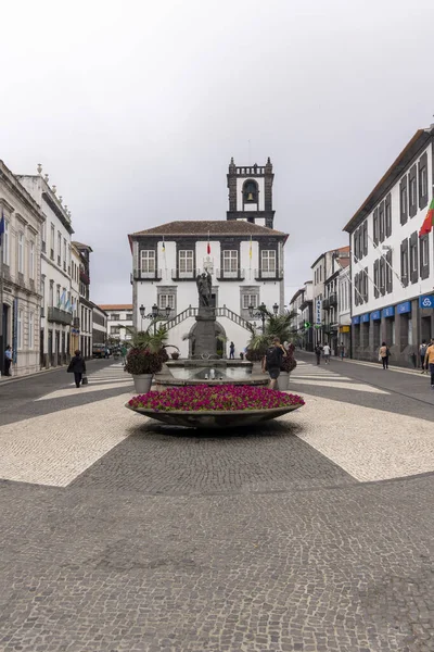 Ponta Delgada, Portekiz, 8 Ekim 2025: Ortasında çeşme, arkasında saat kulesi olan bir sokak sahnesi. Çeşme çiçeklerle çevrili ve birkaç kişi etrafta dolaşıyor.