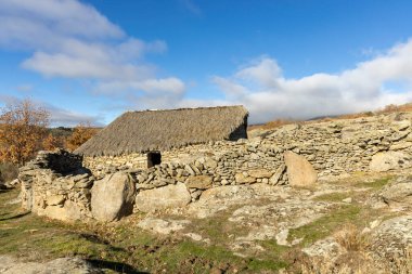 Yamaçta küçük, yıpranmış bir taş bina var. Binanın etrafı kayalarla çevrili ve sazdan bir çatısı var. Gökyüzü açık ve mavidir, ve güneş parlıyor. Huzurlu bir sahne.