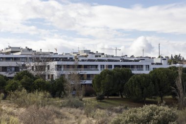 Önünde bir sürü penceresi ve yeşil ağacı olan büyük bir bina. Gökyüzü bulutlu ve güneş görünmüyor.
