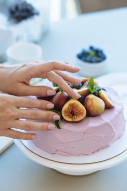 Ön plandaki şekerlemecinin elleri açık pembe sünger pastayı bulanık masa, yemekler ve malzemelerin arka planında meyve, nane ve böğürtlenlerle süsler