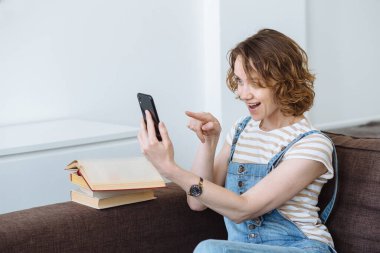 Genç neşeli kız öğrenci oturma odasında kanepede oturuyor ve canlı çevrimiçi diyaloglar kuruyor. Video çağrısı ile uzak iletişim.