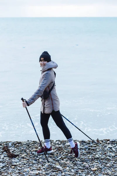 Young sports woman walks along rocky coast of sea bay on Scandinavian sticks. Healthy lifestyle, physical activity.