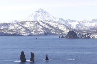 Russia, Kamchatka Peninsula. Three brothers rocks in the beautiful water of the bay and the Vilyuchinsky volcano in the background. Untouched nature of Russia. Suitable for postcards and advertising. clipart