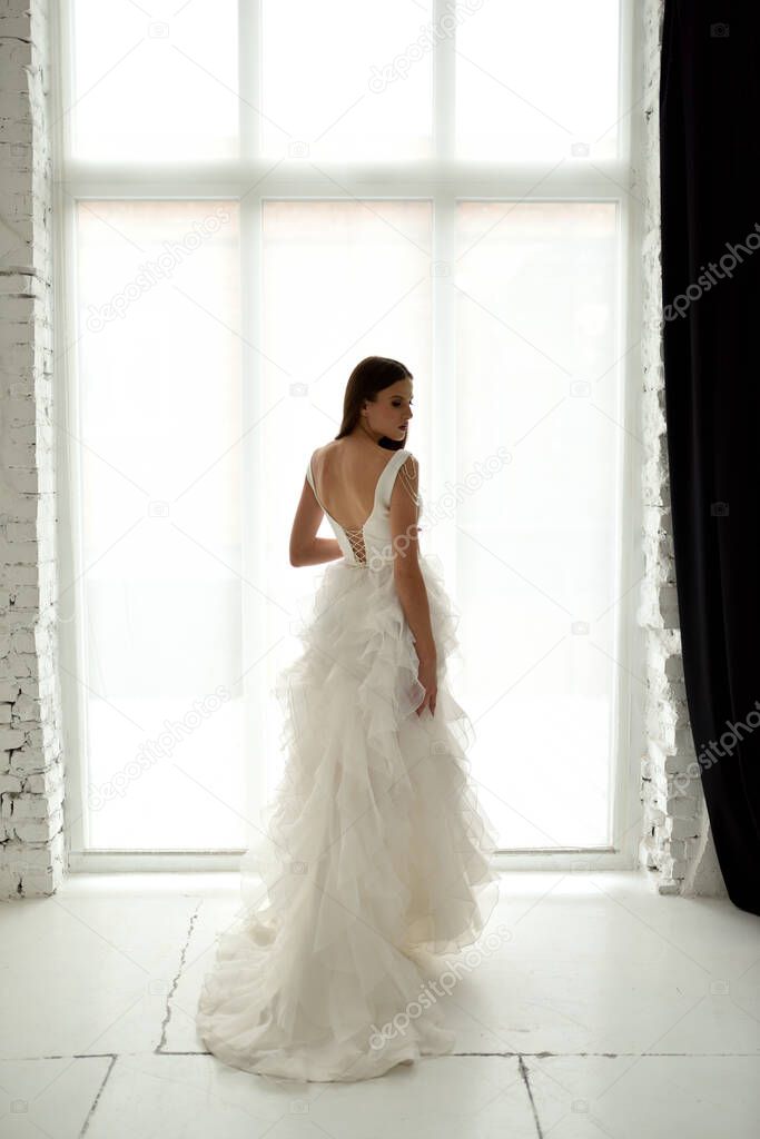 Portrait of a beautiful bride in full growth in the studio on a white background. The woman stands with her back and looks into the frame.
