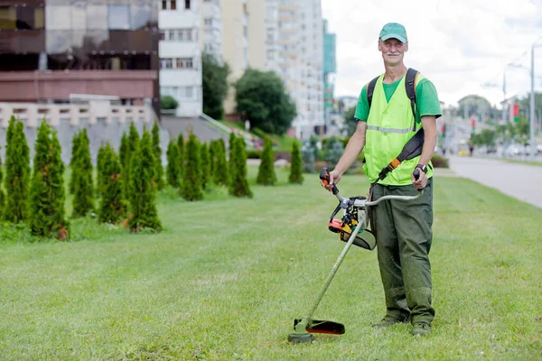 市のサービス都市の通りの背景に芝生のムーバー草のトリマーとシニア労働者の庭師 ロイヤリティフリーのストック写真