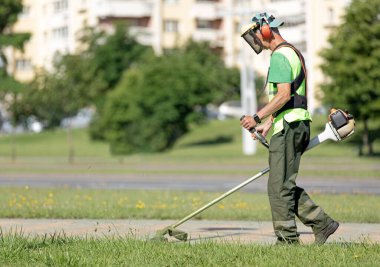 Municipal Gardener Landscaper Man Worker Cutting Grass with String Trimmer Machine along City Street clipart
